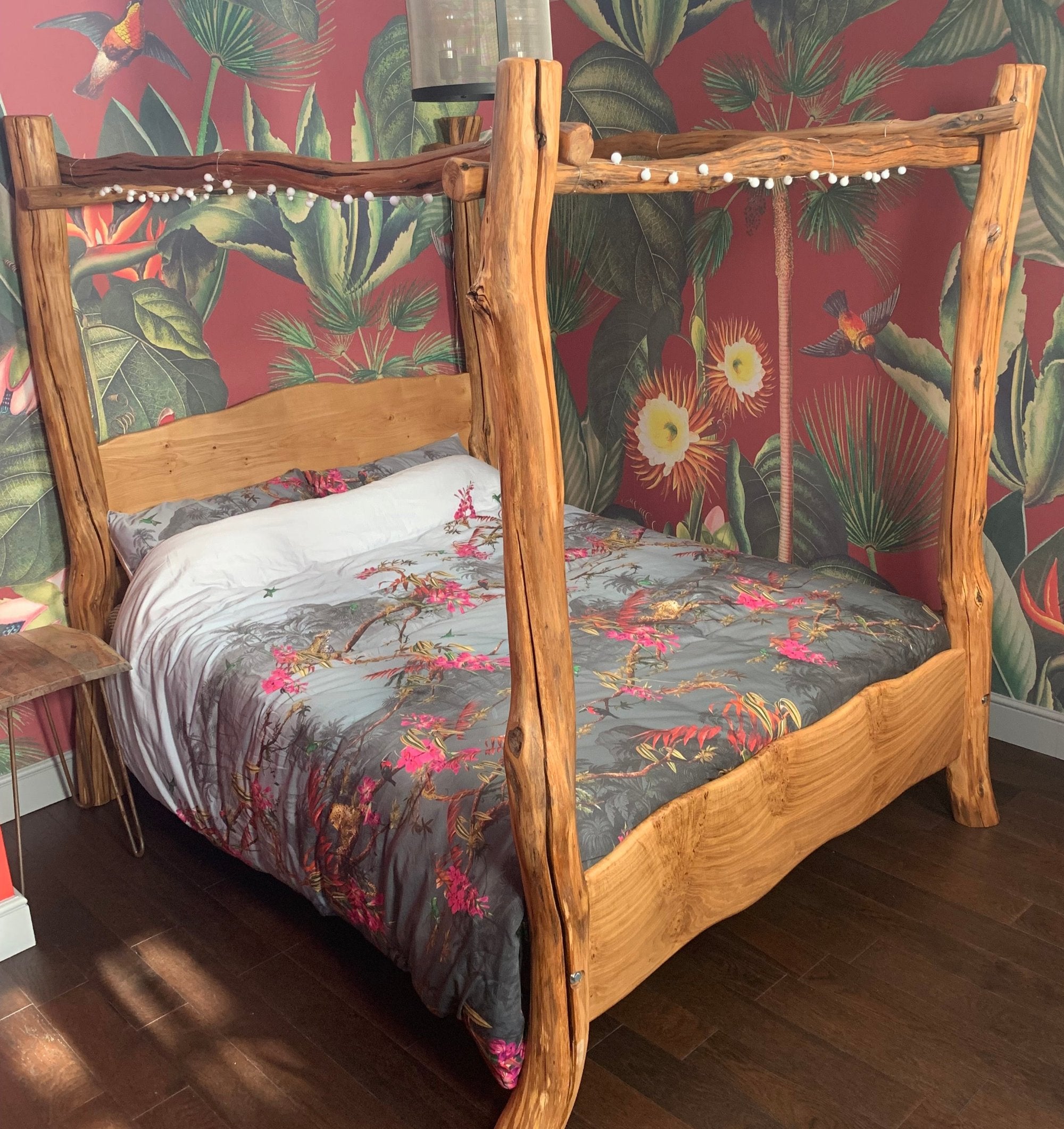 Rustic four-poster bed frame in natural wood with unique grain details, styled with floral bedding and bold botanical wallpaper, creating a vibrant and cozy bedroom for Janine