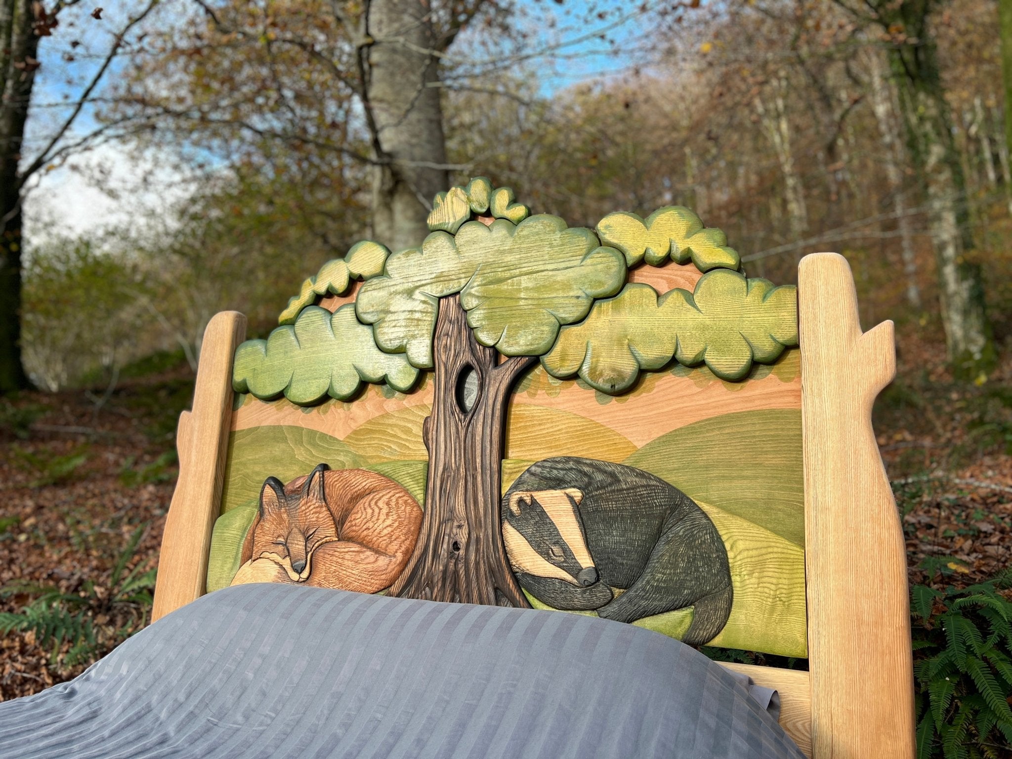 Close-up view of a hand-carved wooden bed headboard featuring a detailed depiction of a sleeping fox and a curled-up badger under tree branches, showcasing intricate craftsmanship and a nature-inspired design.