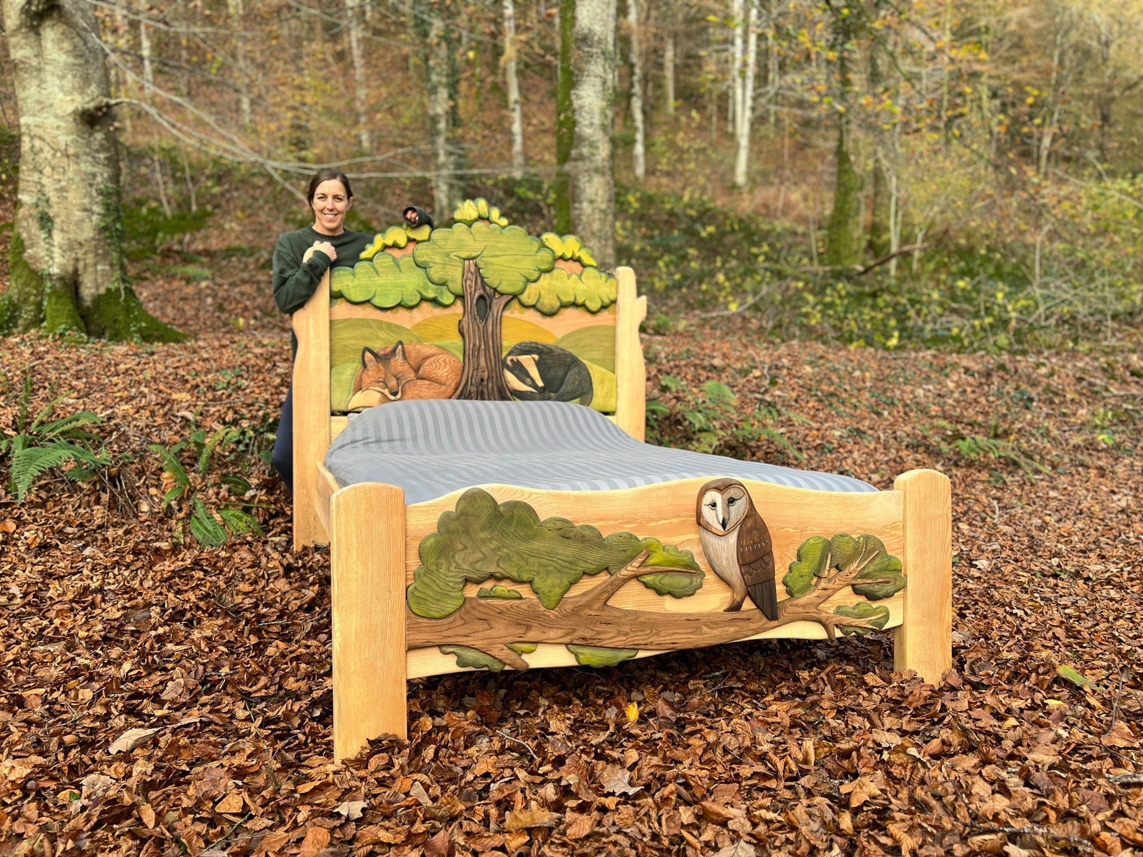 A woman standing beside a hand-carved, nature-inspired wooden bed featuring detailed carvings of a fox, badger, and owl, set in a forest backdrop. The bed showcases intricate craftsmanship and is made of solid wood, highlighting a unique blend of art and functionality.