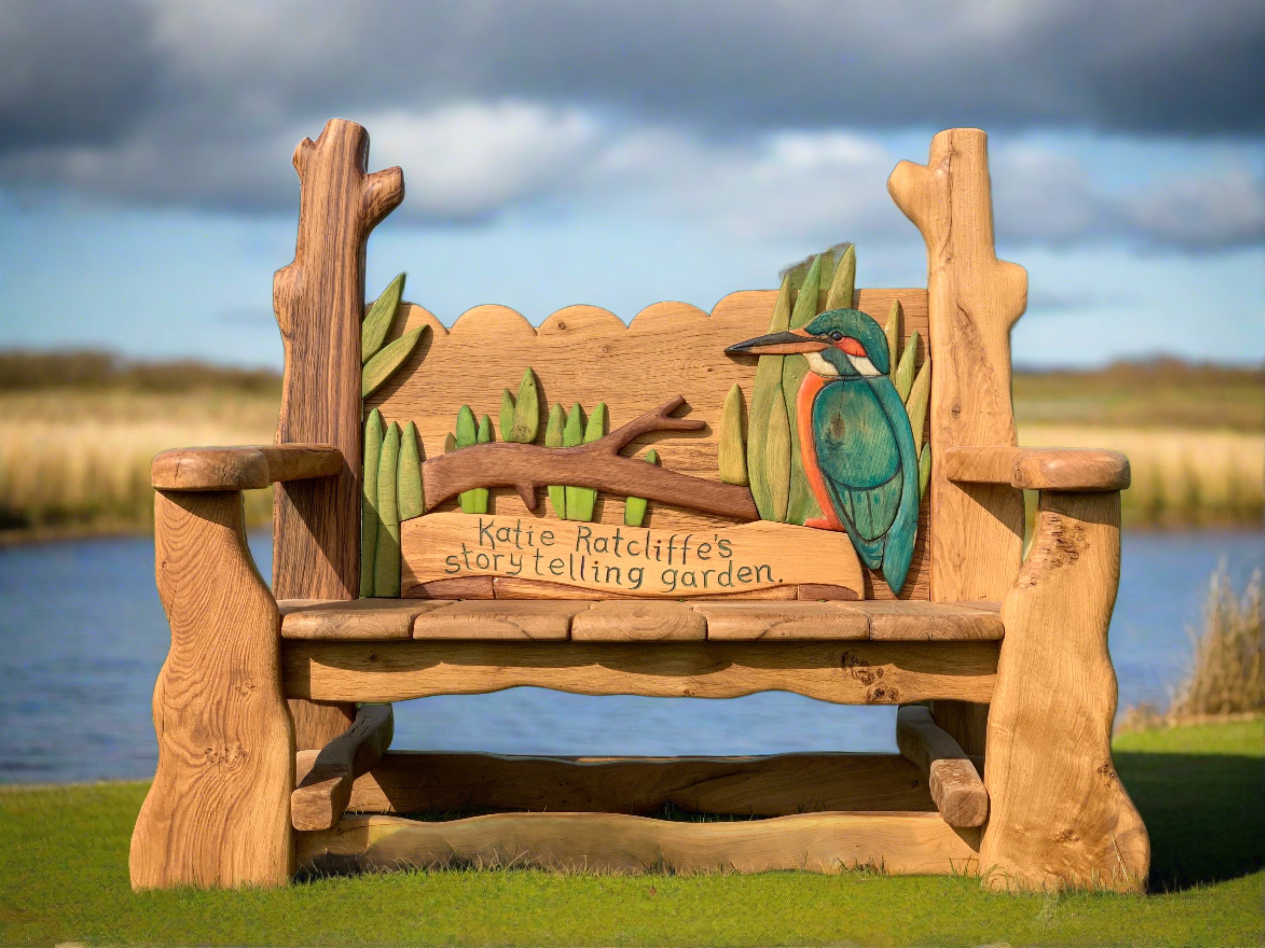 Banc commémoratif en bois fabriqué à la main par Free Range Designs, orné d'un martin-pêcheur et de roseaux magnifiquement sculptés, dédié au jardin des contes de Katie Ratcliffe, offrant un espace tranquille et artistique pour se souvenir et raconter des histoires.