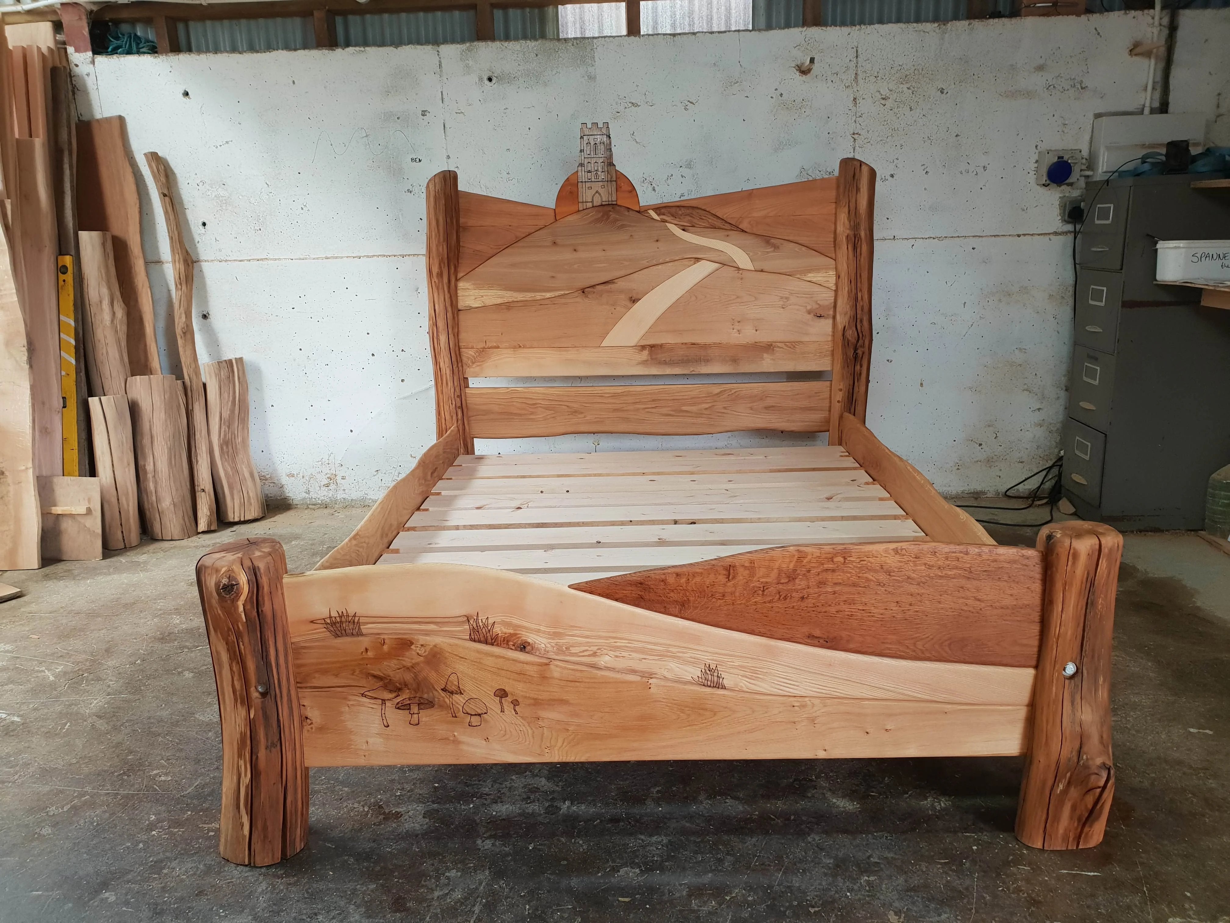 Close-up of the Glastonbury Tor bed frame in a workshop, showing the detailed carving of the Tor and winding path on the headboard