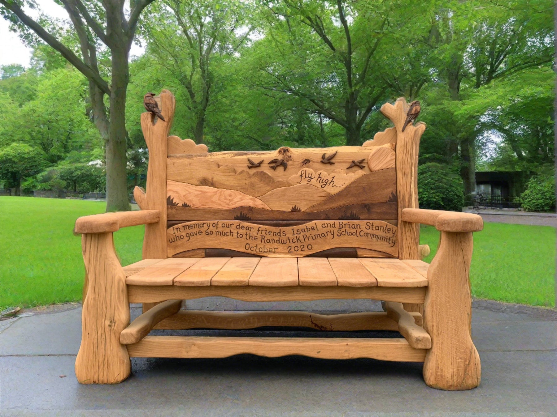 Handcrafted memorial bench featuring bird carvings and a scenic landscape, inscribed with the message 'In memory of our dear friends Isabel and Brian Stanley' at Randwick Primary School. The backrest displays the phrase 'Fly high' with birds soaring in the background.