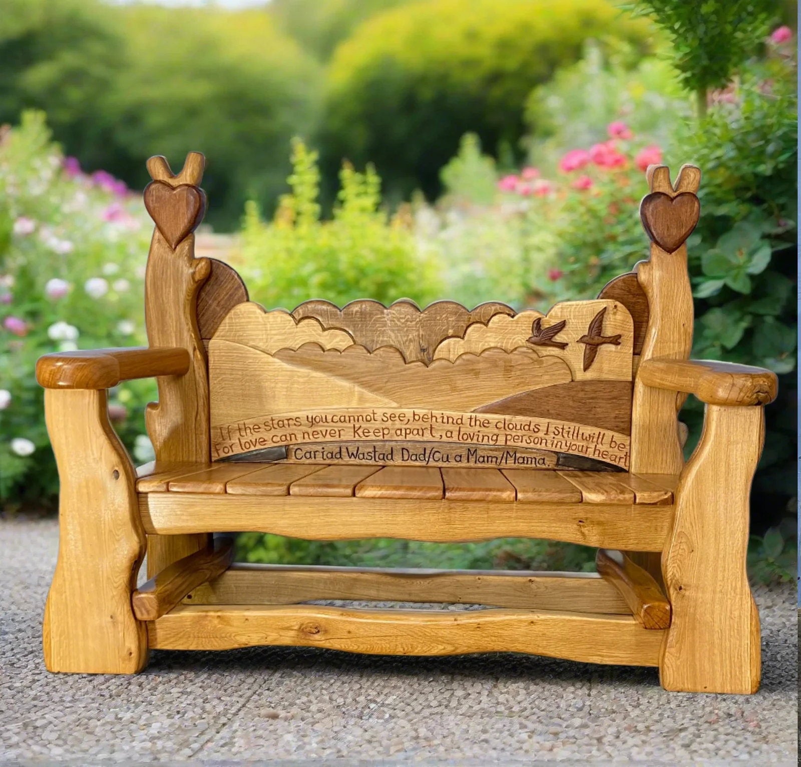 Gedenk-Holzbank mit detaillierten Schnitzereien von Vögeln, Wolken und einer malerischen Landschaft auf der Rückenlehne. Die Bank ist mit einem Tribut an Isabel und Brian Stanley graviert, in einer ruhigen Parklandschaft mit grünen Bäumen im Hintergrund