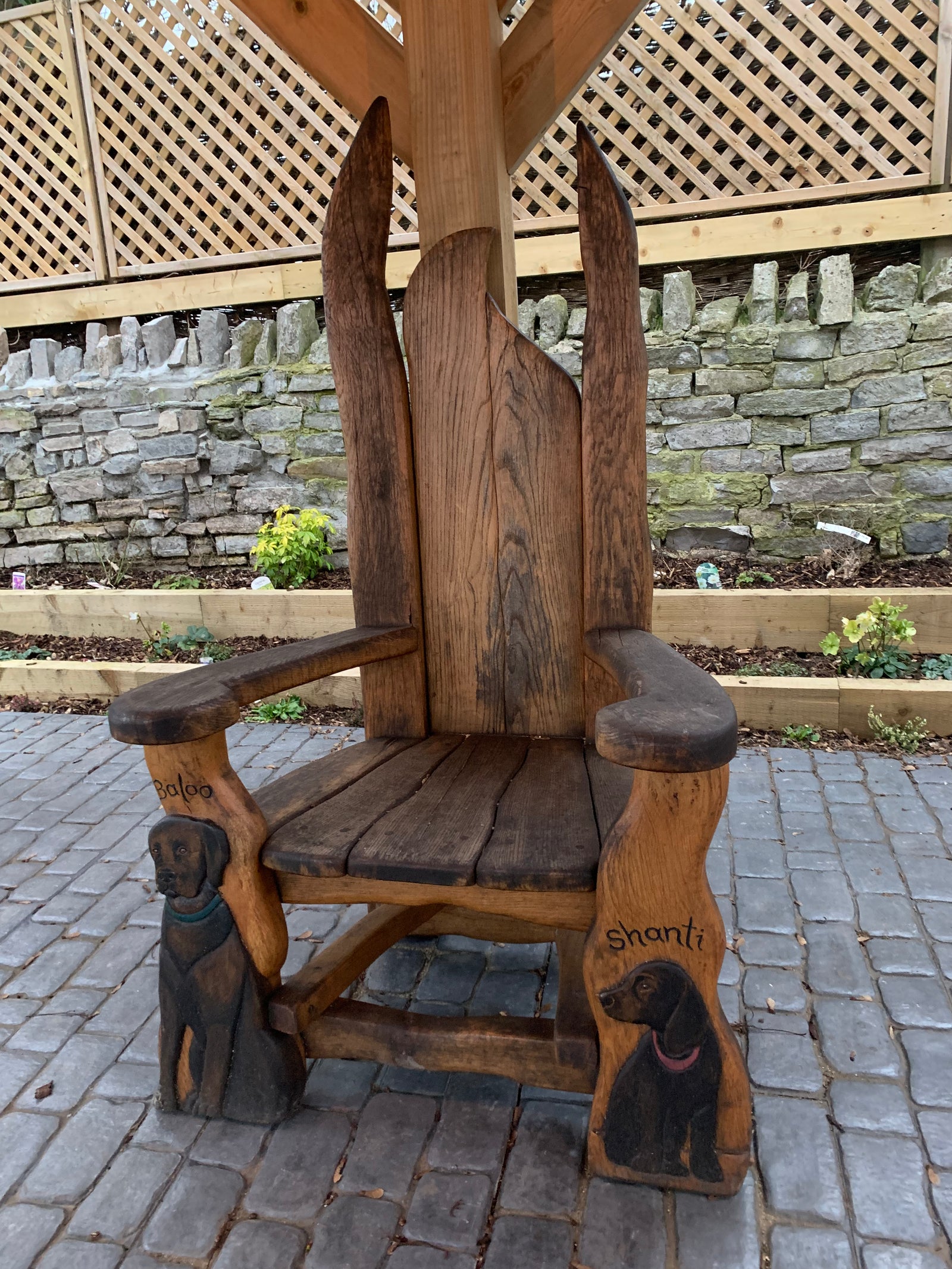 A handcrafted memorial wooden chair with detailed carvings of dogs Baloo and Shanti, placed on a cobblestone patio with a rustic stone and wooden lattice backdrop, blending artistry and remembrance.
