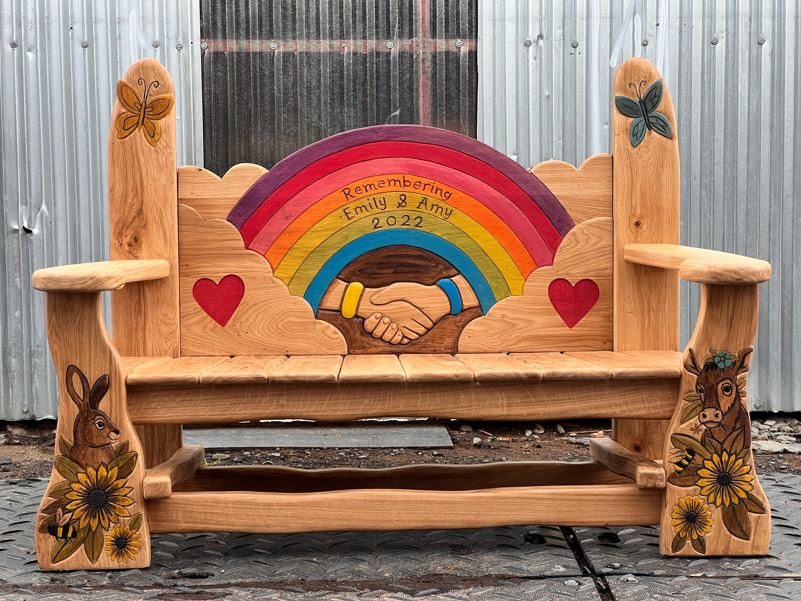 Bespoke hand-carved oak memorial bench front view with rainbow, clasped hands wearing friendship bracelets and red hearts