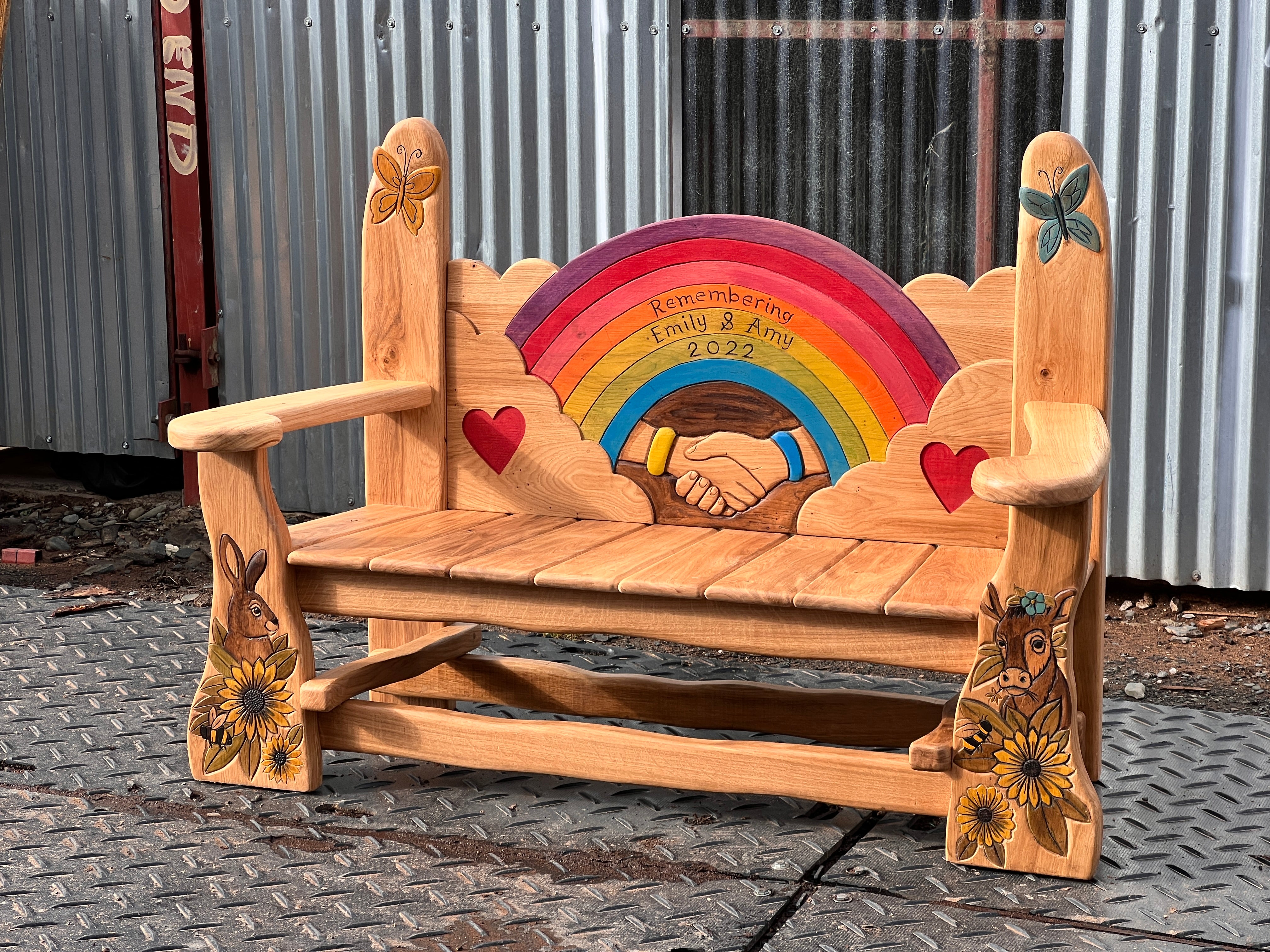Bespoke solid oak memorial bench direct front view showing full rainbow panel, hearts, butterflies and animal carvings