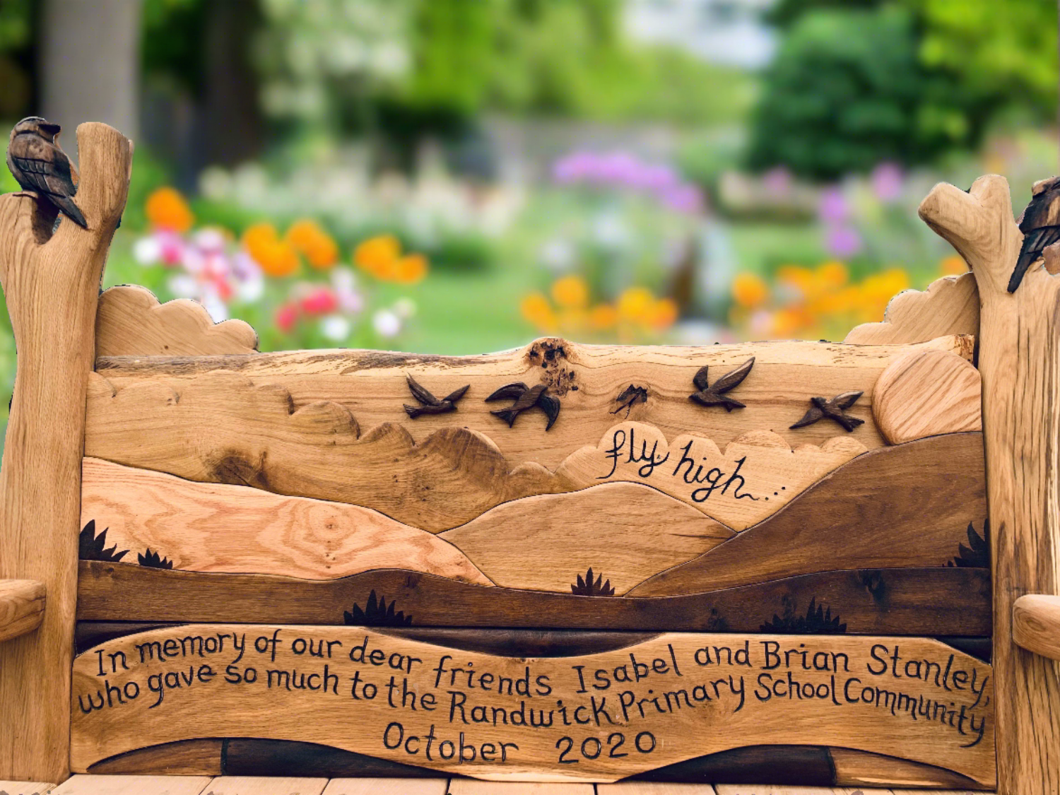 Handcrafted memorial wooden bench with detailed carvings of birds, a scenic landscape, and an engraved message in honor of Isabel and Brian Stanley, set in a tranquil park