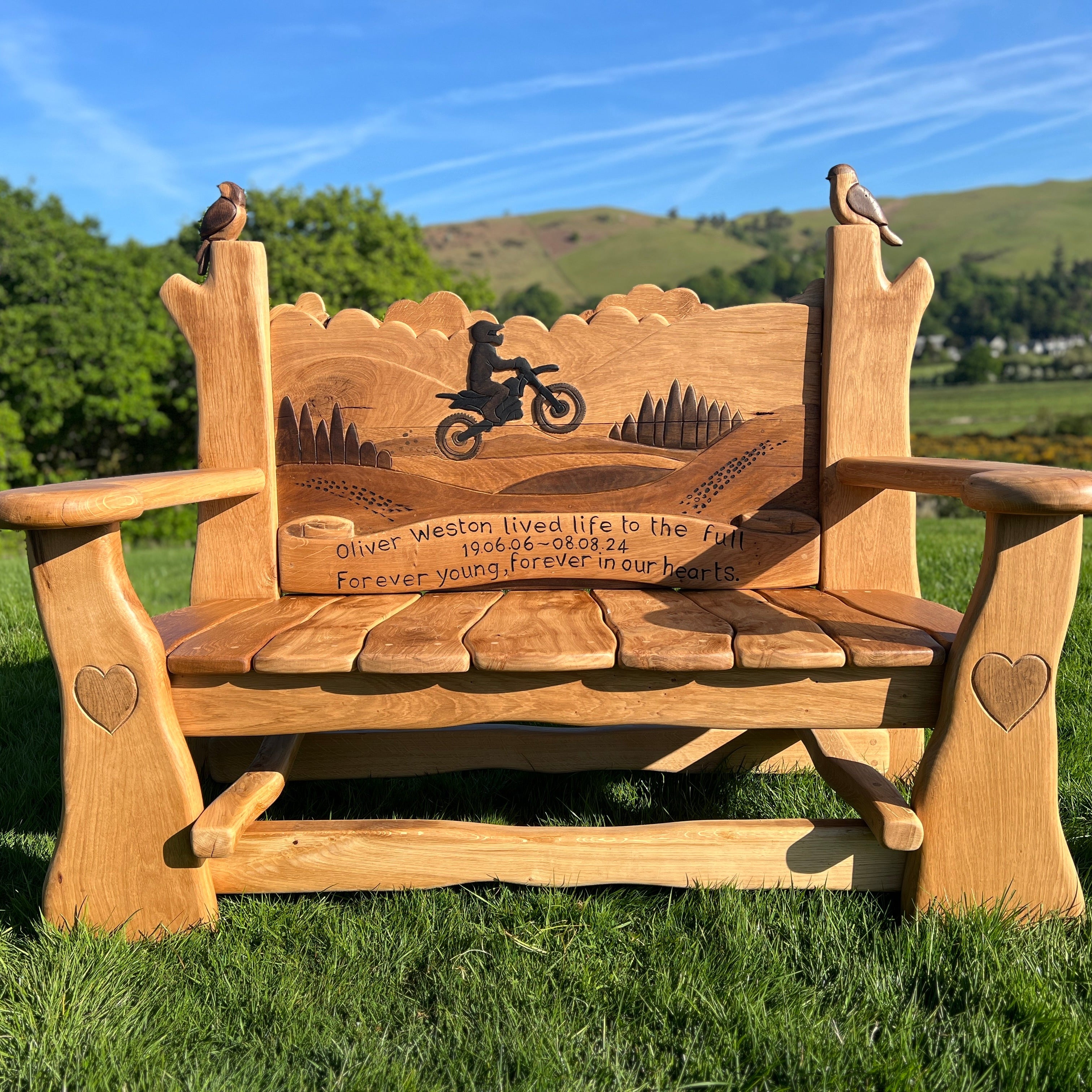 Hand-carved oak memorial bench with motorbike rider silhouette and engraved tribute to Oliver Weston, set in a grassy landscape with countryside background.

