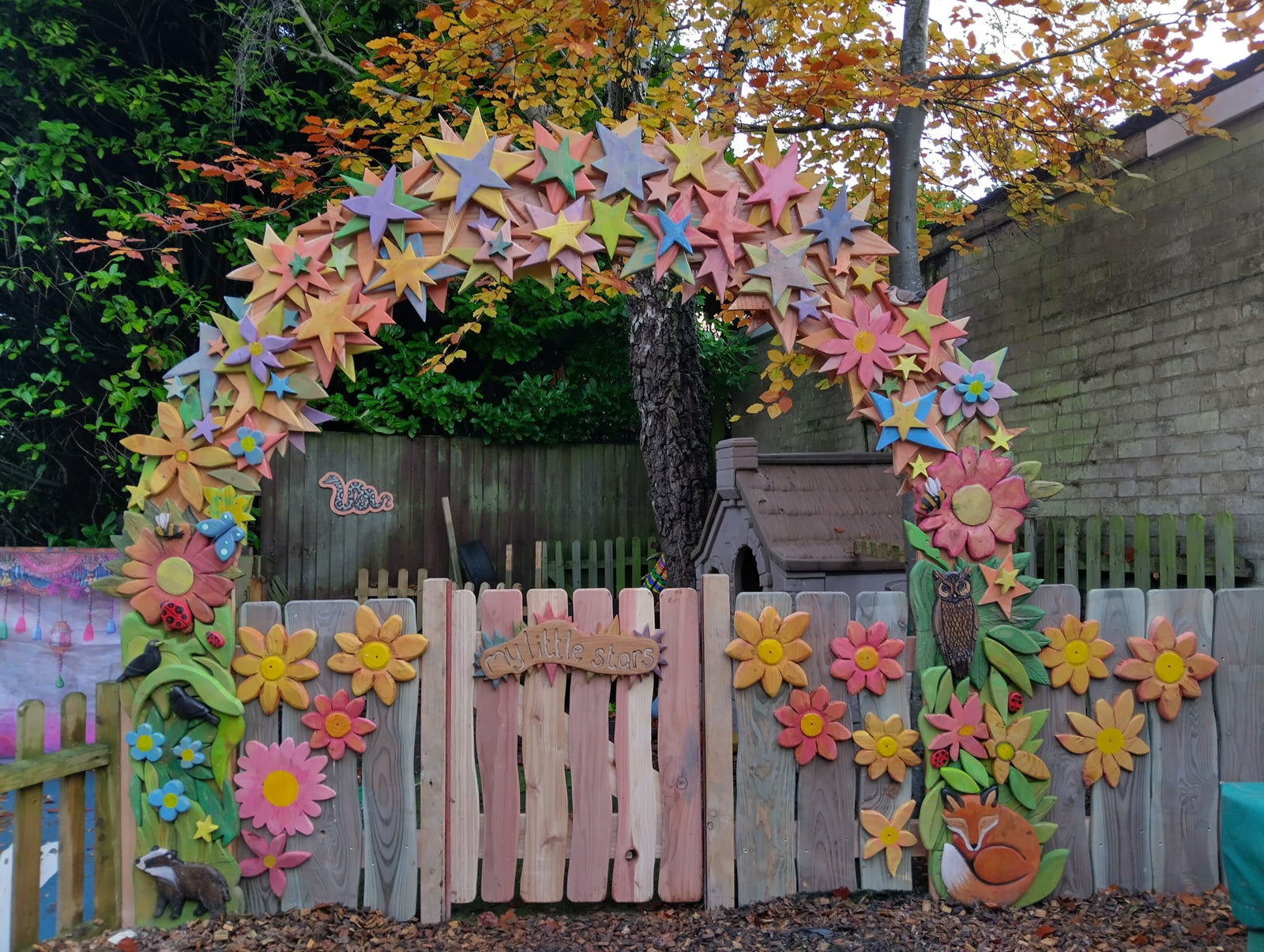 Decorative arch with colorful flowers and stars above a wooden fence in a backyard setting.
