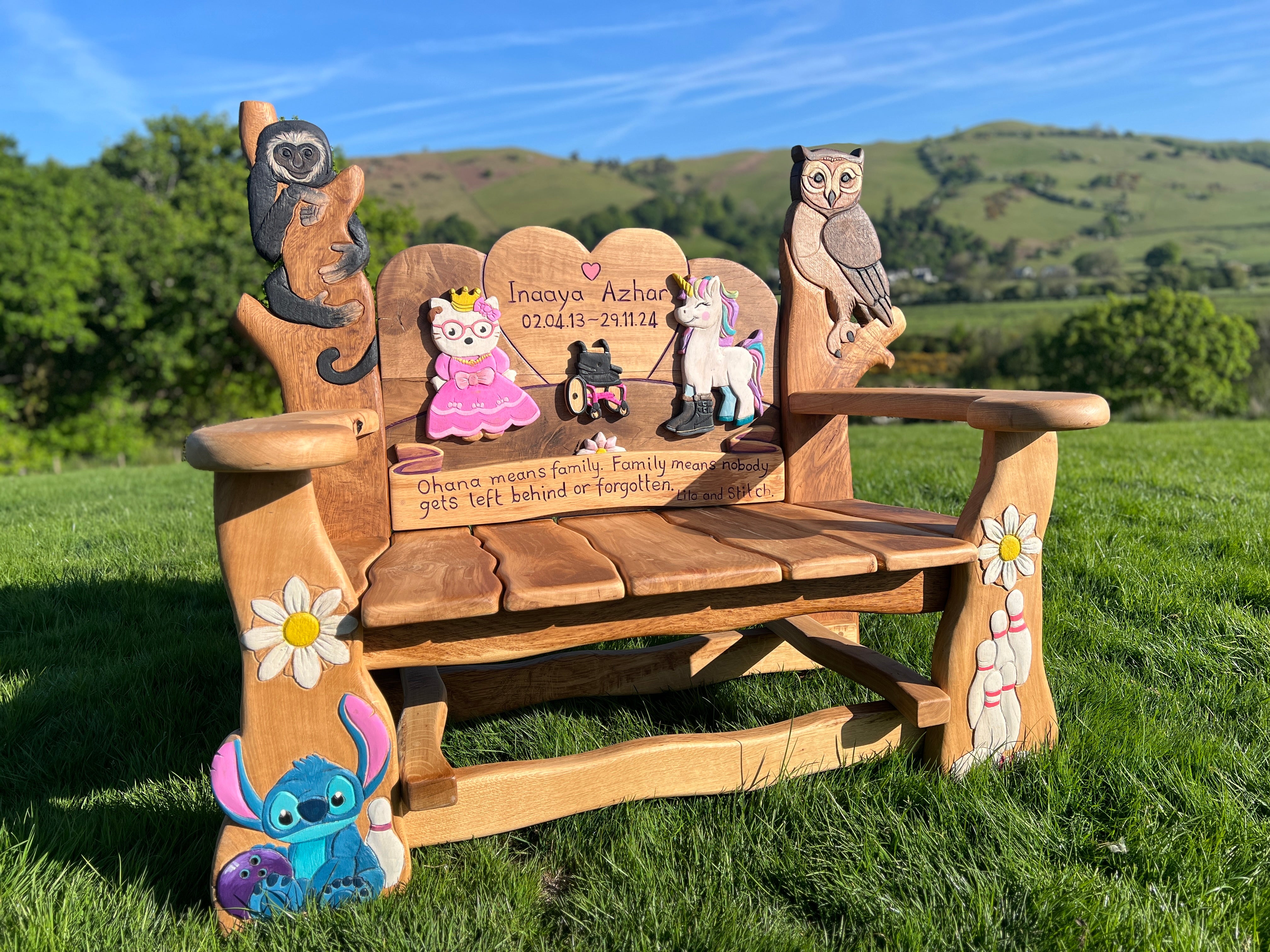 Angled view of colourful custom children’s memorial bench with carved animals and cartoon characters in a natural oak setting.
