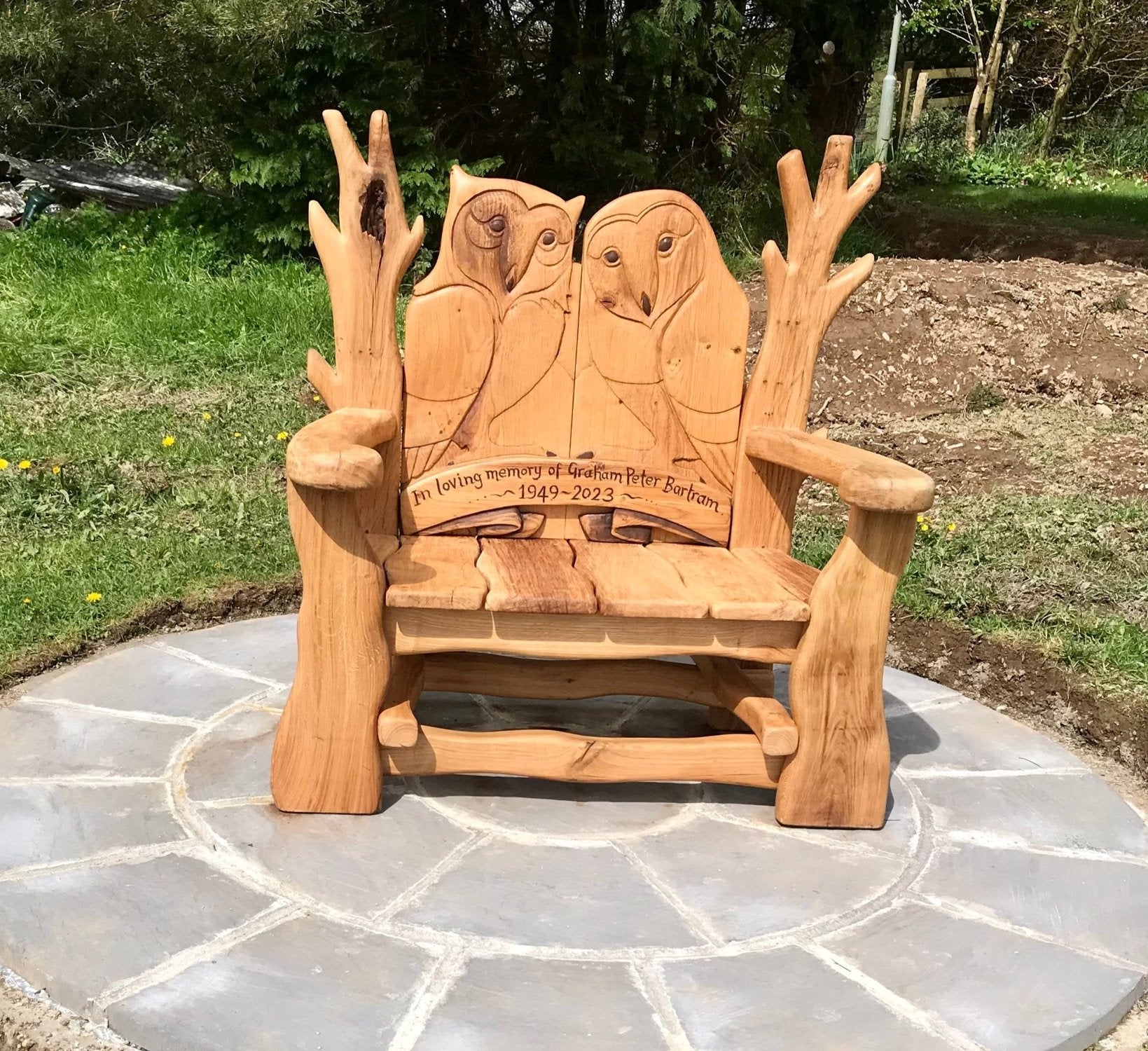 Handcrafted wooden memorial bench with carved owl design, placed on a stone patio in a natural garden setting