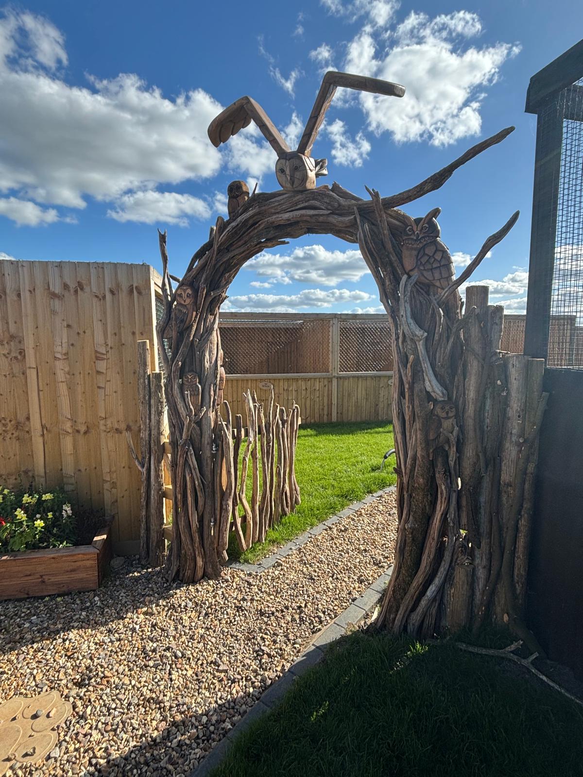 Owl arch installed at an aviary entrance with a rustic wooden gate beneath a blue sky.