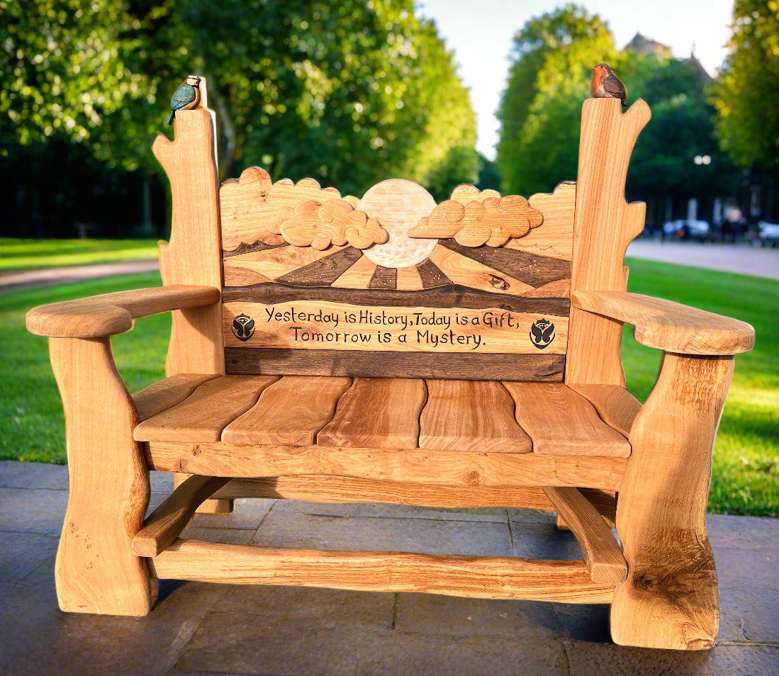 "Handcrafted wooden memorial bench with a detailed carved backrest depicting a sunrise, rolling hills, and clouds. The bench is engraved with the quote: ‘Yesterday is History, Today is a Gift, Tomorrow is a Mystery.’ A robin and a blue tit are perched on the backrest, symbolizing remembrance and connection to nature. The bench is set in a sunlit park with a tree-lined avenue in the background, creating a tranquil and reflective setting