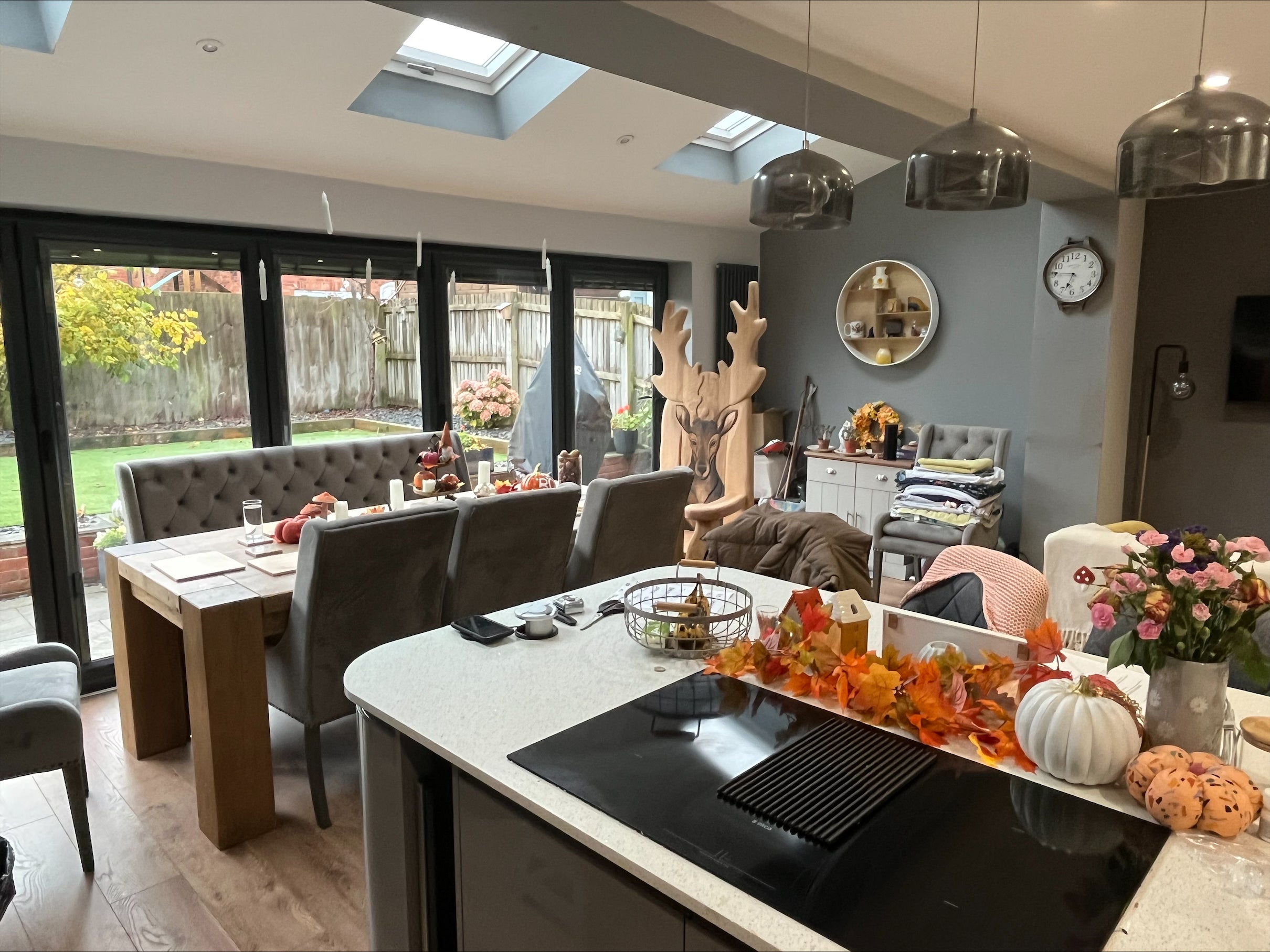 A cozy, modern kitchen and dining area decorated for autumn with colorful leaves and pumpkins. The dining table is set, and in the background, a unique wooden chair with antler-style design adds a rustic touch