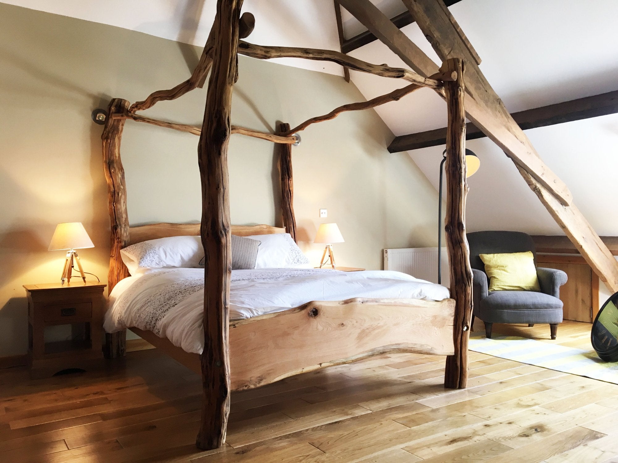 Magnifique lit à baldaquin en chêne fabriqué à la main avec des éléments en bois naturel, placé dans une chambre mansardée confortable avec des planchers en bois et un éclairage doux, offrant une ambiance rustique mais élégante.