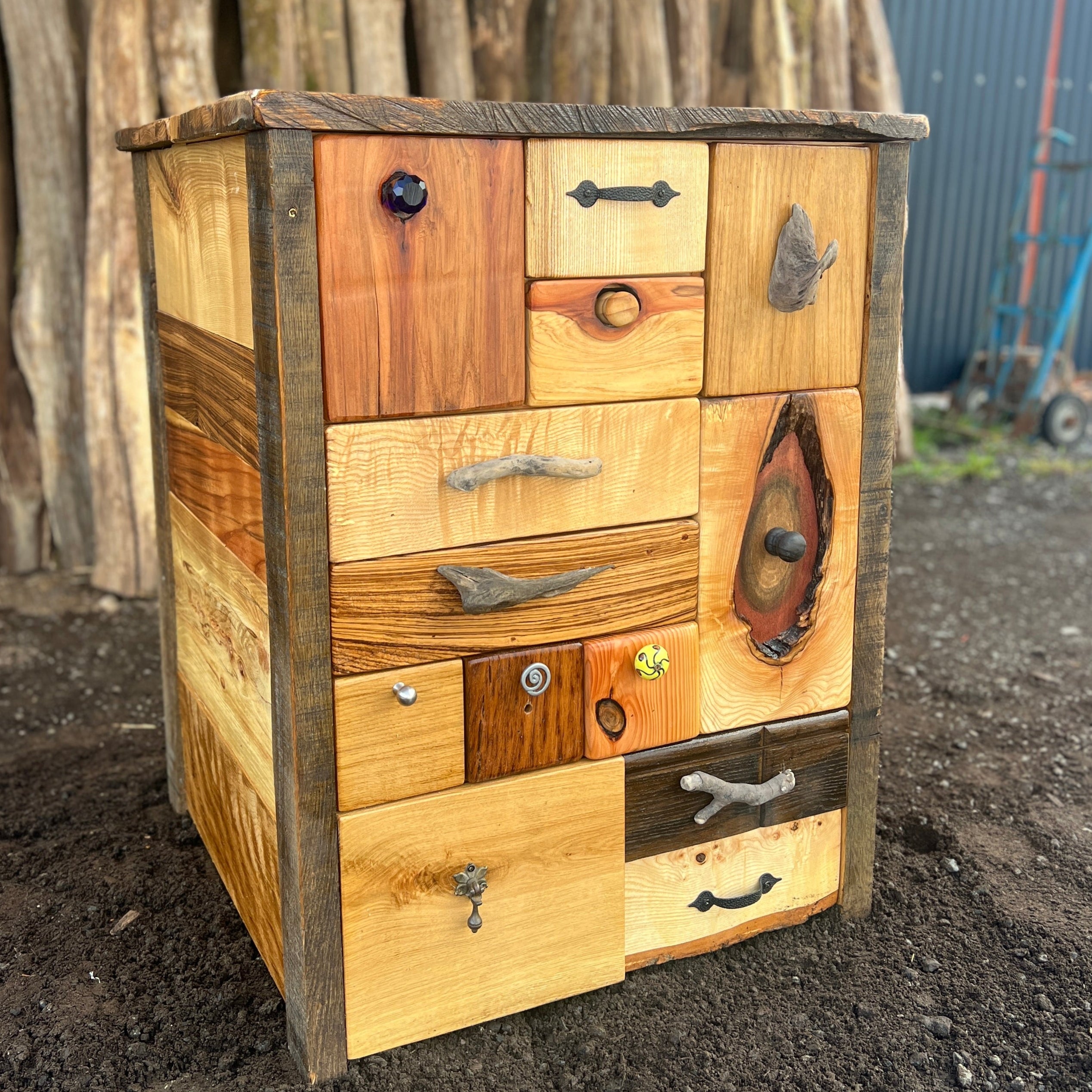 Wooden dresser with various wooden drawers against a backdrop of stacked logs.