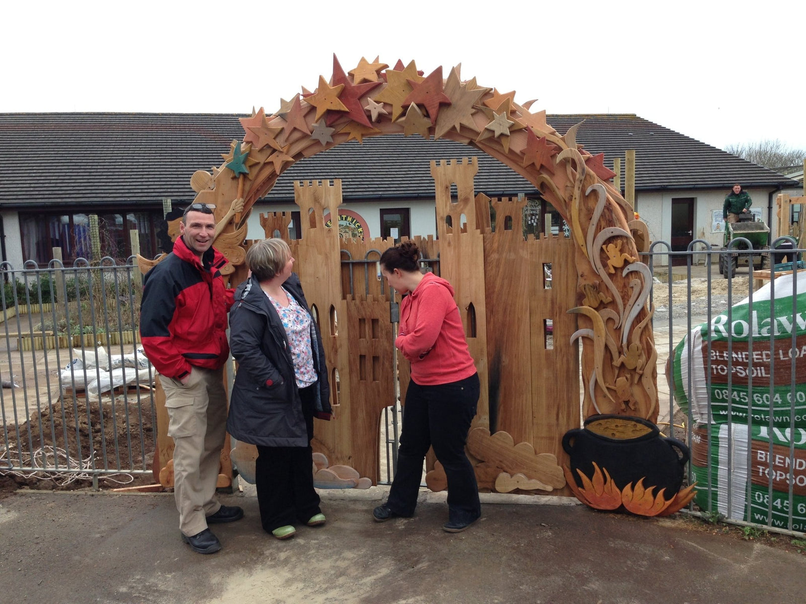 Three people standing in front of a large wooden arch with star designs, near a garden center.