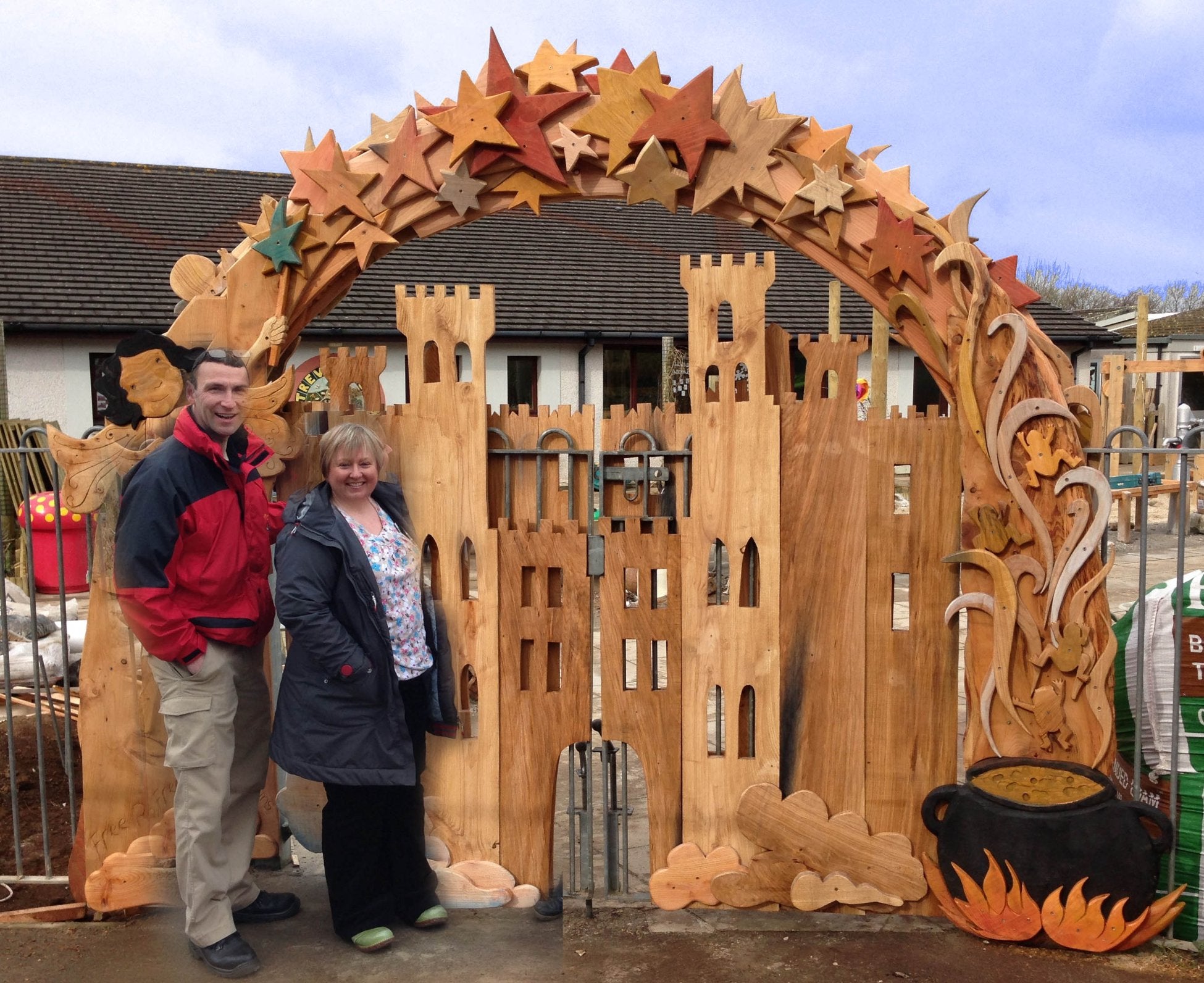 Two people standing beside a large, intricately carved wooden gate featuring a castle, a cauldron with flames, and an arch of colorful stars above, showcasing whimsical and artistic woodworking.