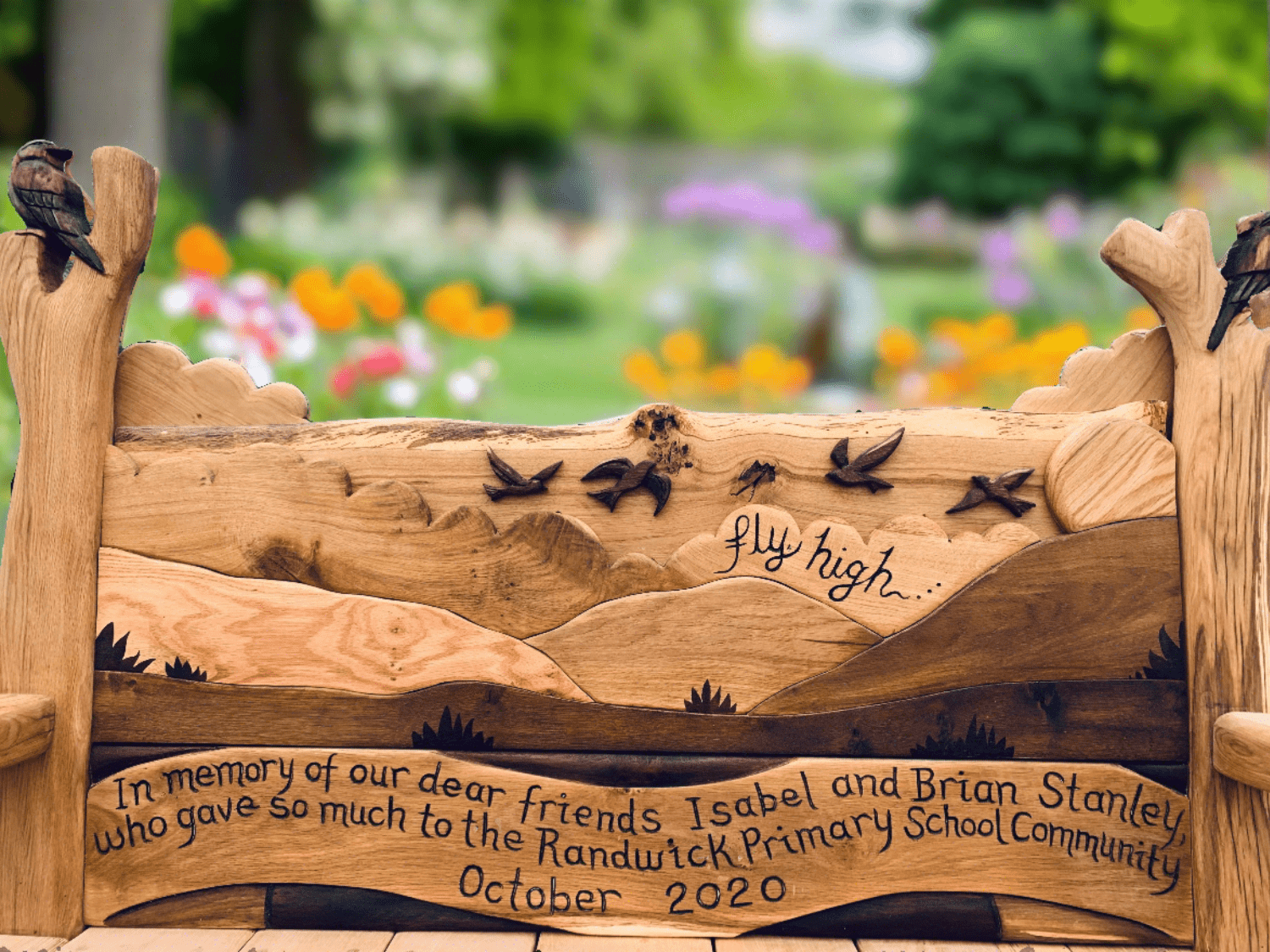 Handcrafted memorial wooden bench with detailed carvings of birds, a scenic landscape, and an engraved message in honor of Isabel and Brian Stanley, set in a tranquil park