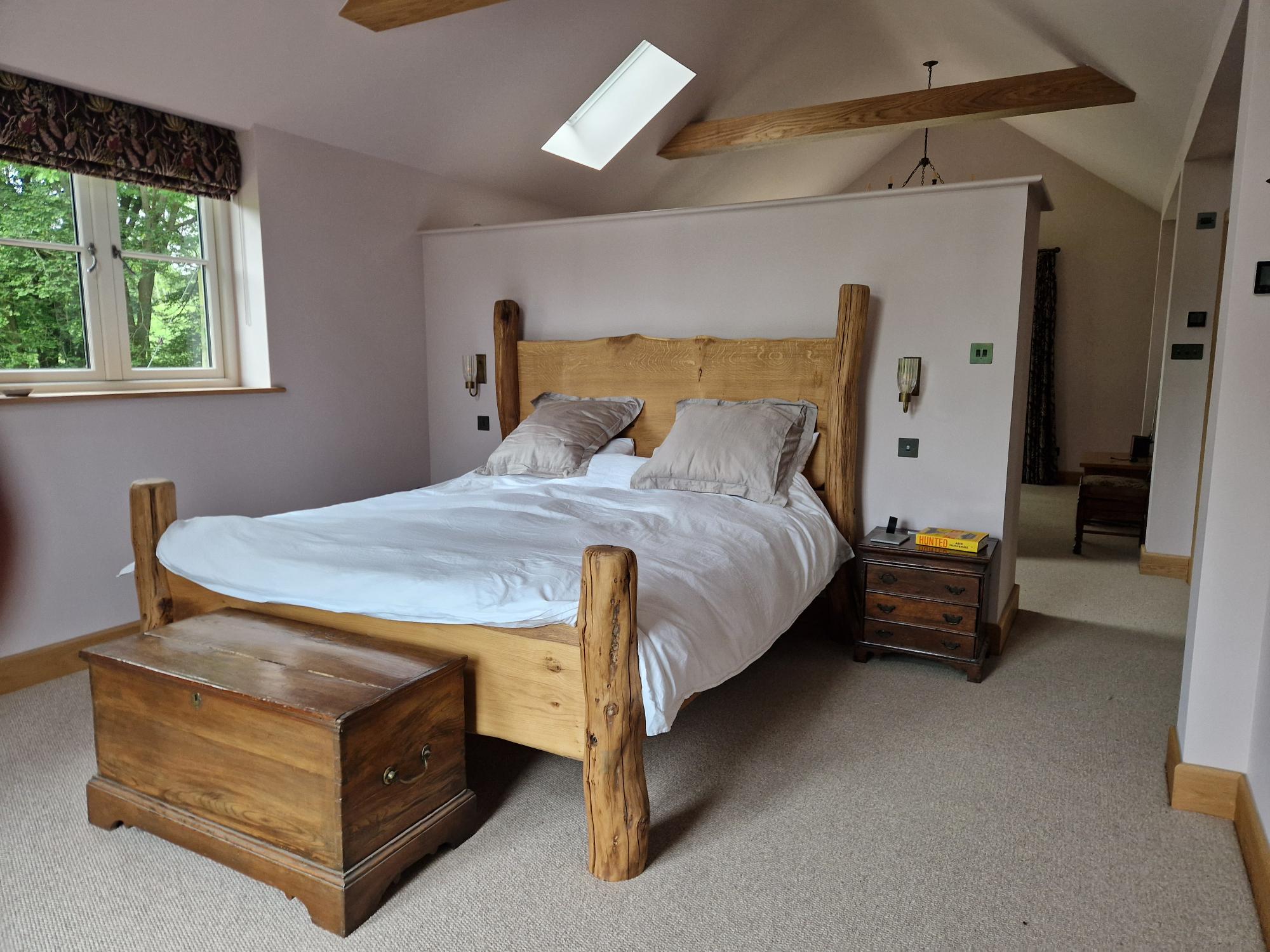 Lit artisanal en chêne massif au design rustique, installé dans une chambre spacieuse baignée de lumière naturelle. Le lit se caractérise par des montants en bois robustes et une tête de lit en bois lisse, complétés par un coffre en bois au pied du lit.