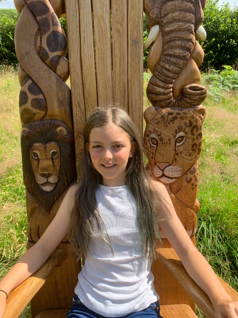 Young girl sitting on a beautifully carved storytelling chair with intricate animal designs, including a lion, leopard, and elephant, crafted by Free Range Designs