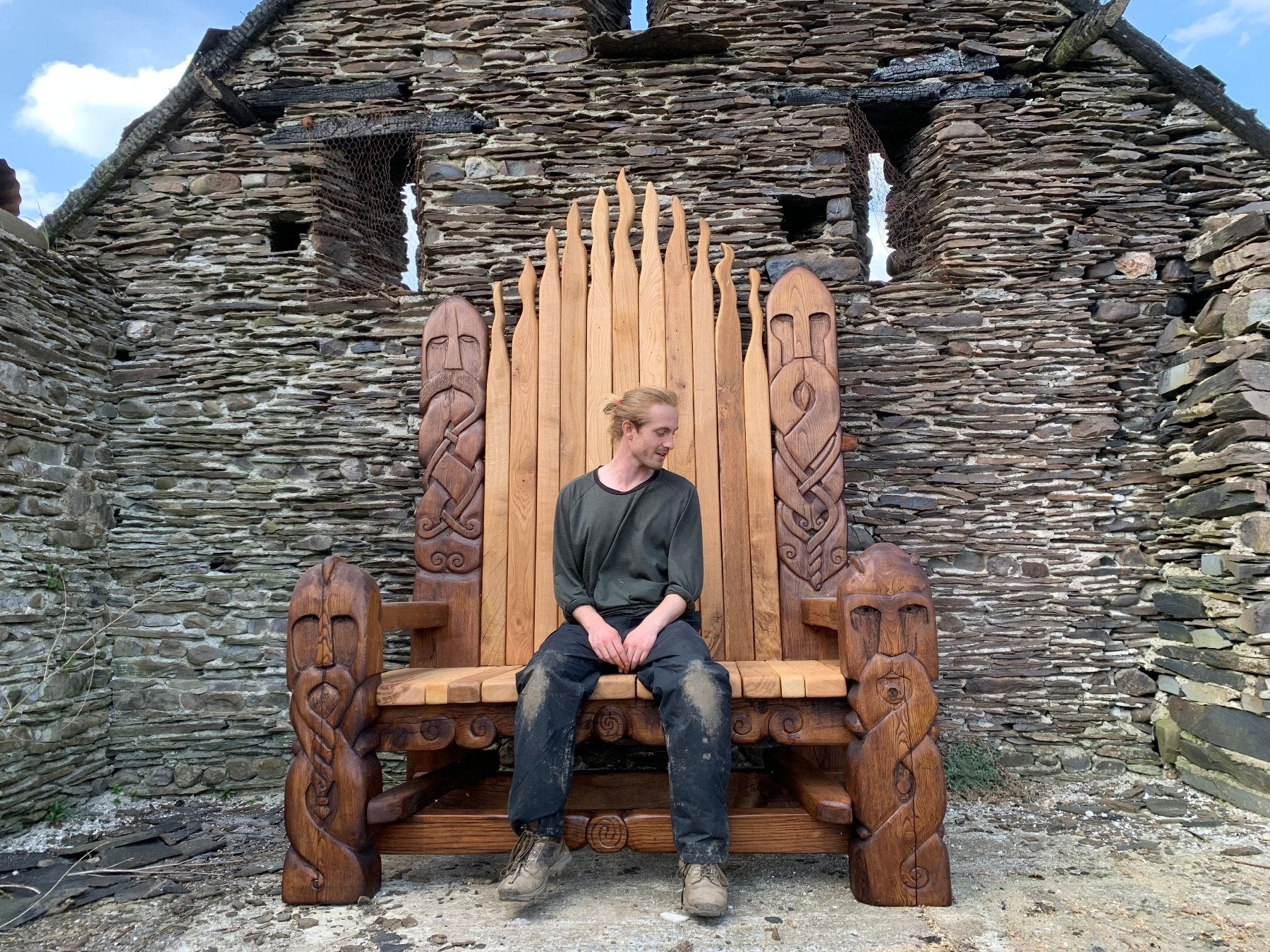 Handcrafted Viking throne chair with intricate Celtic carvings, featuring a craftsman seated in front of an ancient stone building