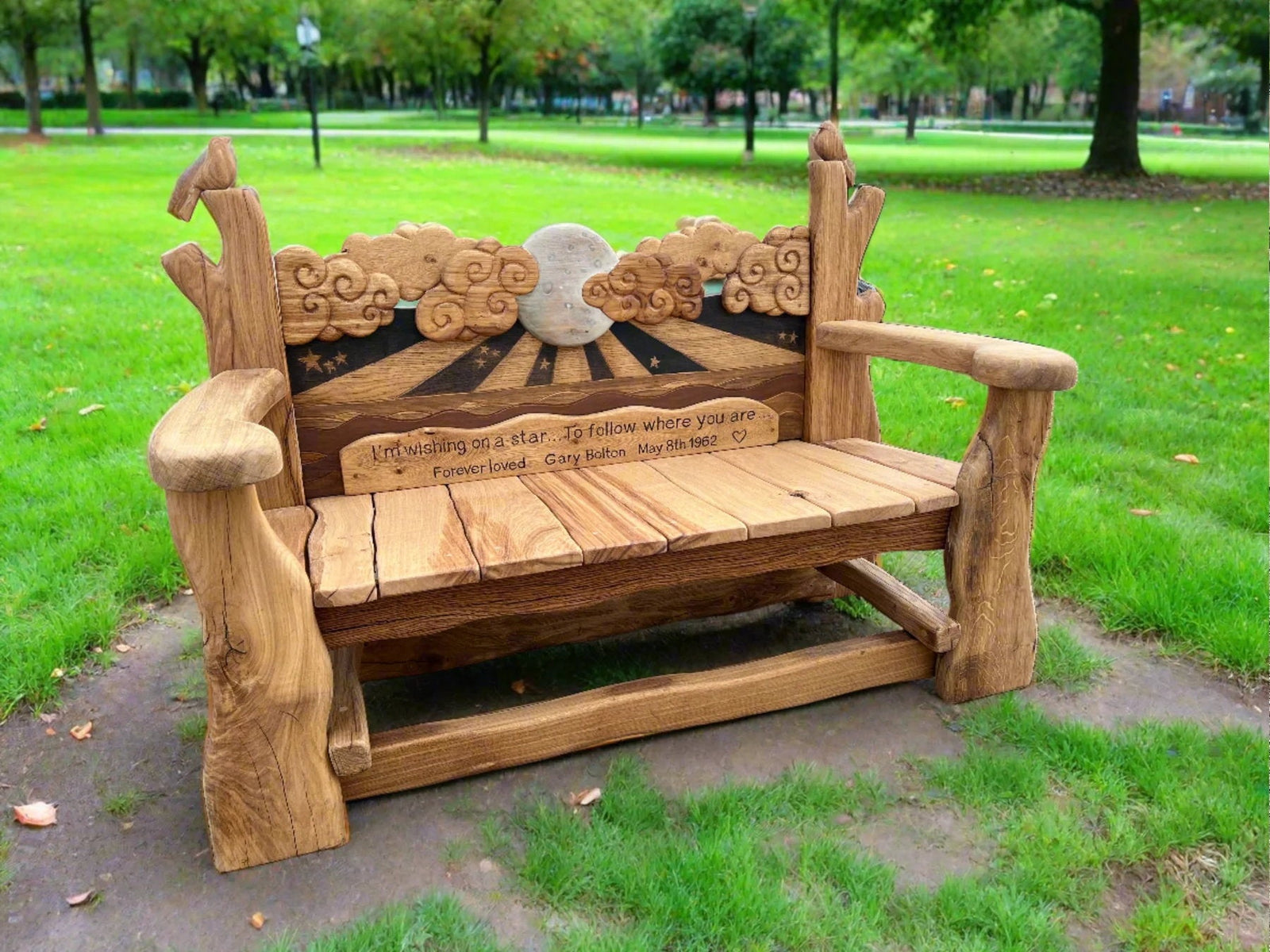 Handcrafted memorial bench in honor of Gary Bolton, showcasing a celestial theme with carved clouds, a luminous moon, and stars. The backrest features the inscription: 'I’m wishing on a star... To follow where you are. Forever loved Gary Bolton, May 8th, 1962.' Placed in a peaceful park, the bench is adorned with bird carvings, symbolizing love, memory, and tranquility
