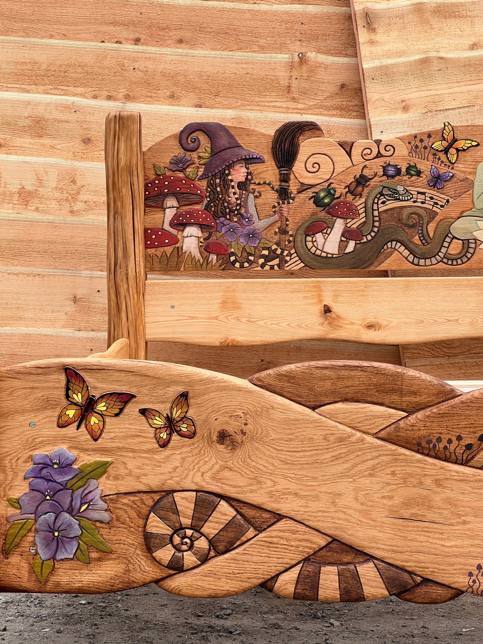 Close-up of hand-carved witch figure among red fly agaric mushrooms and purple flowers on oak bed headboard