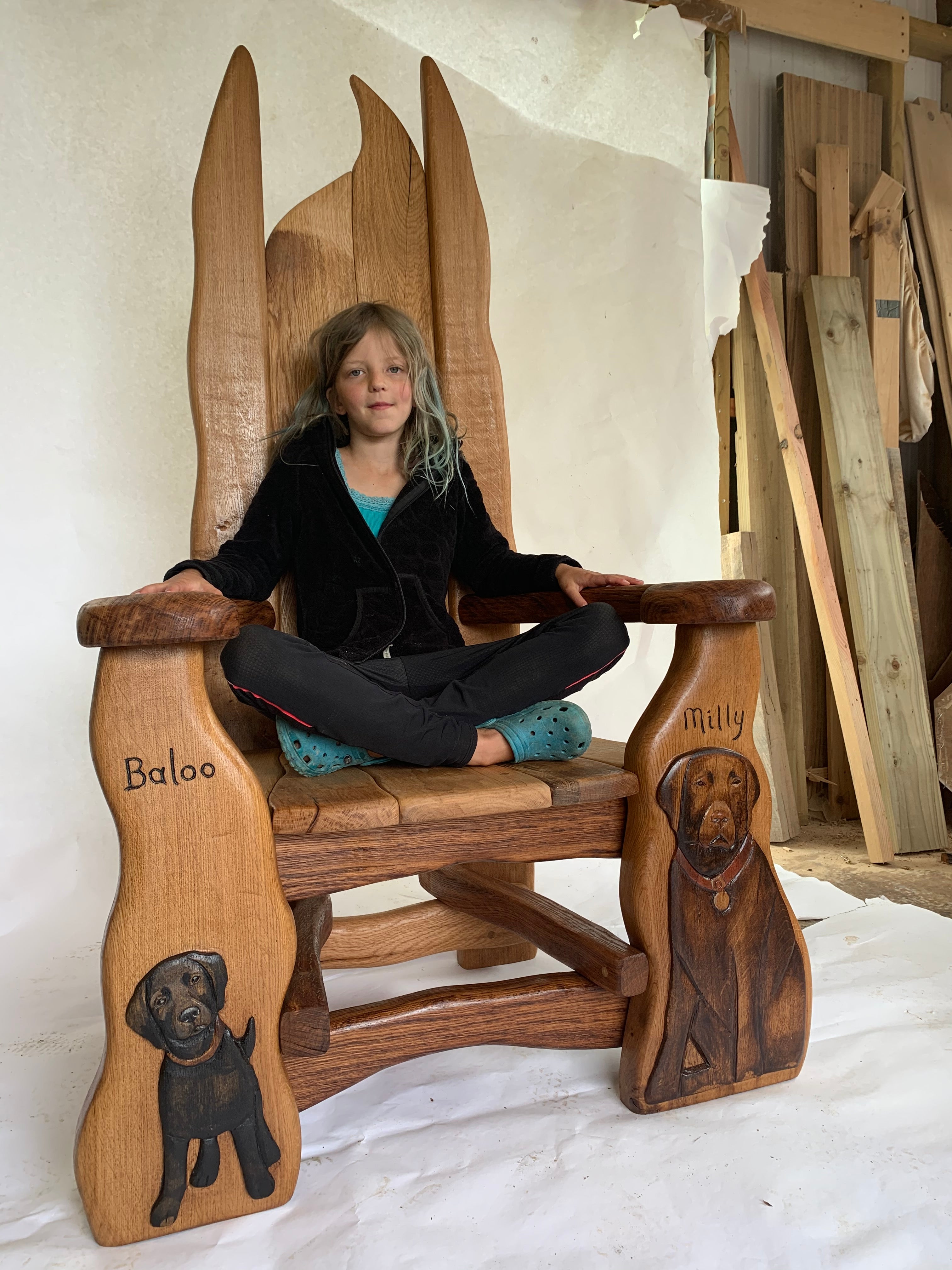 A handcrafted wooden chair with unique carvings of dogs named Baloo and Milly, featuring a young girl seated cross-legged, highlighting the chair's intricate craftsmanship and comfort.