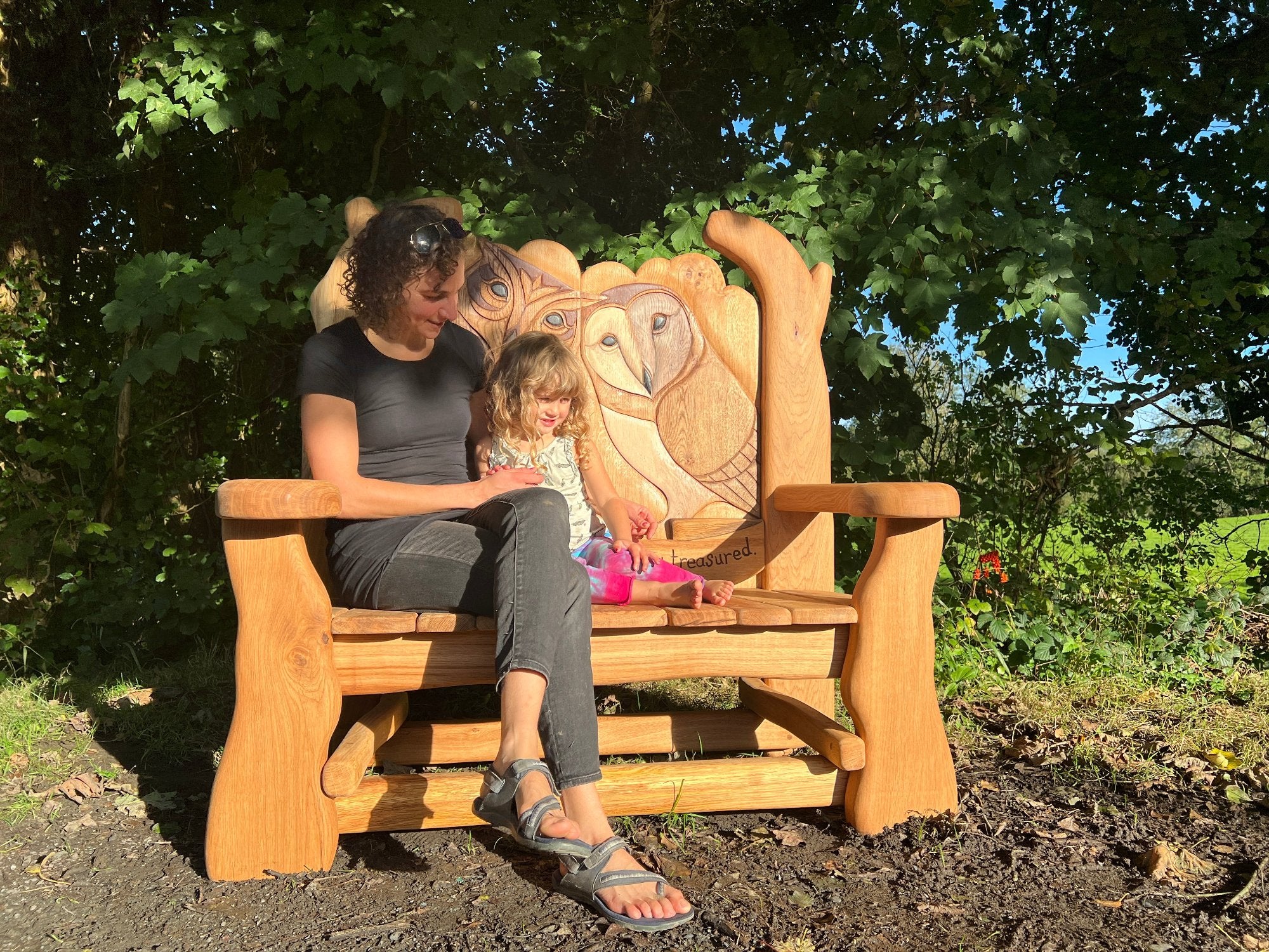 A mother and daughter sitting on a beautifully carved wooden memorial bench featuring detailed owl designs, enjoying a peaceful moment in nature